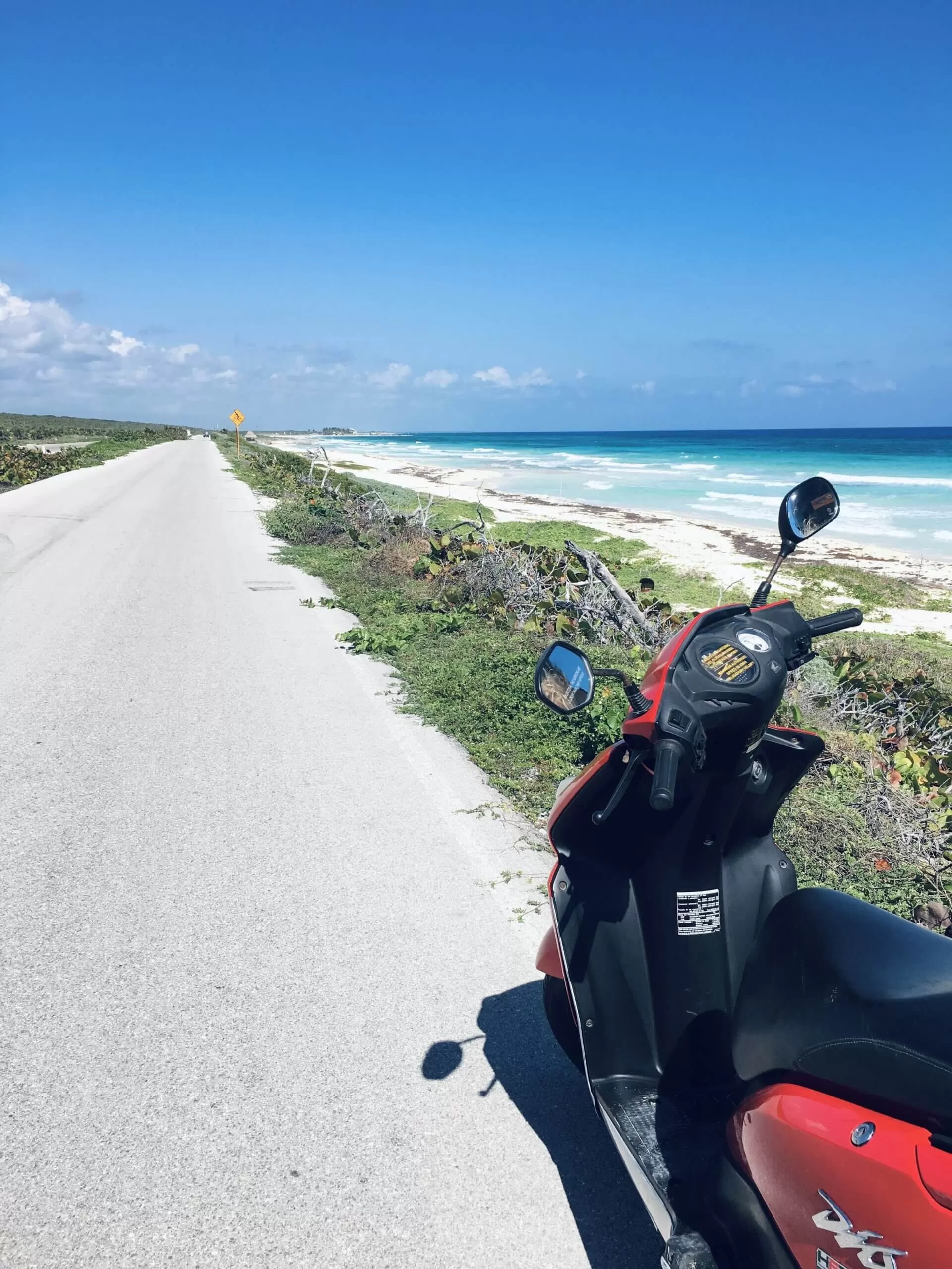 se ve una moto estacionada al lado de la ruta. atras se ve en mar y vegeteacion . esto es en la isla de cozumel, en mexico