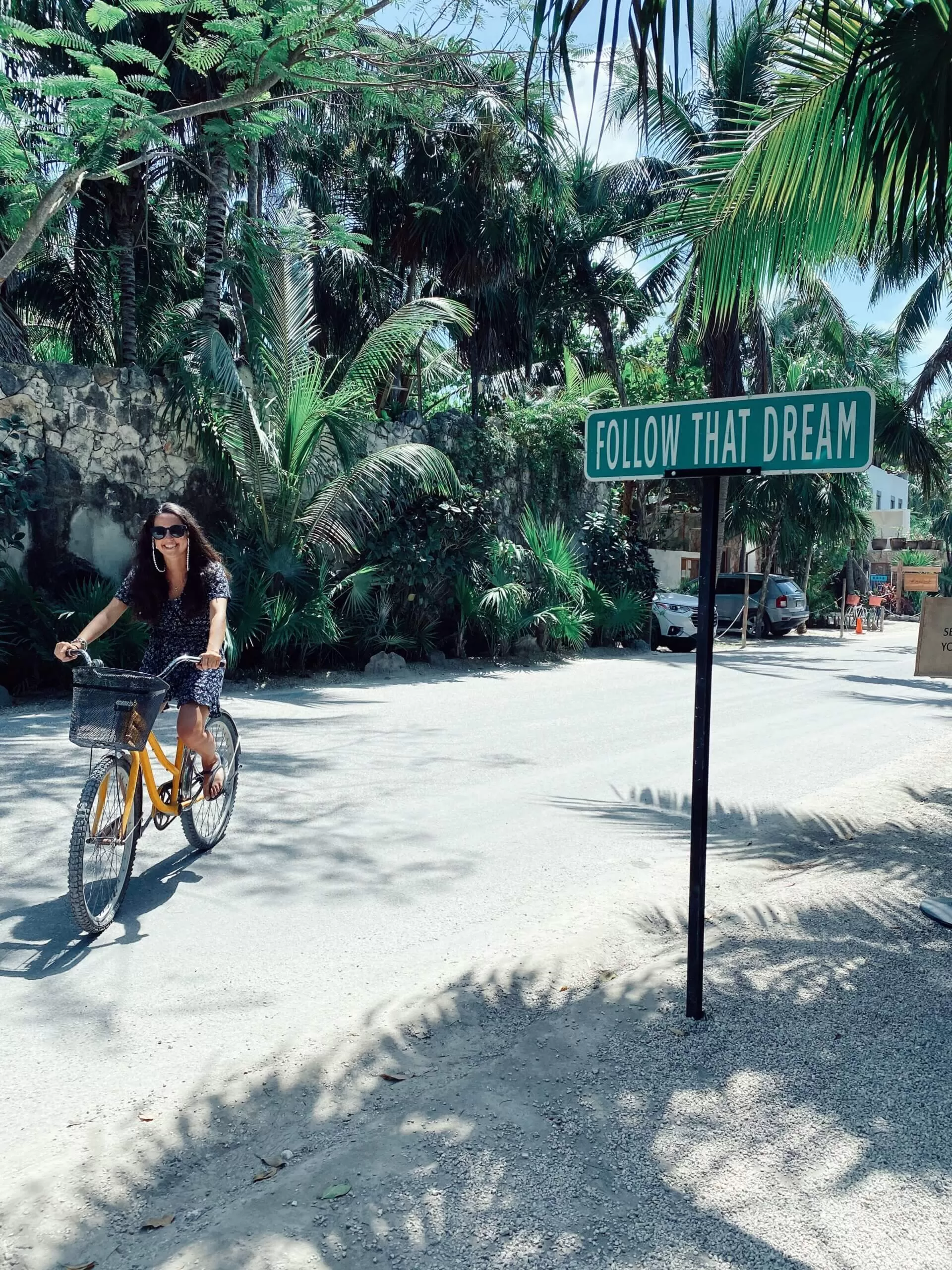 estoy yo andando en bici por las calles de tulum. en la zona hotelera. hay palmeras alrededor y un cartel que dice "follow that dream"