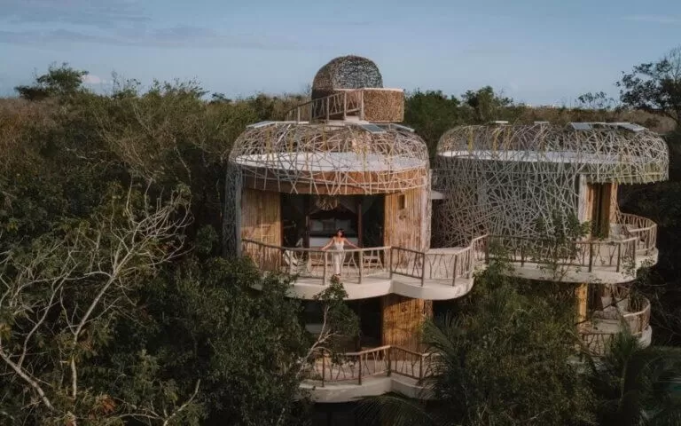 la foto pertenece al hotel kan tulum. es un foto sacada con drone. se puede ver el alojamiento metido en la selva. en un balcon hay una mujer mirando a la selva. el alojamiento tiene una arquitectura particular hecha con lianas de la selva.