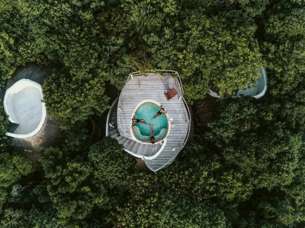 la foto pertenece a Papapaya Playa Project - la foto es tomada con drone desde arriba. se puede ver una pileta en forma de circulo en el techo de una de las habitaciones. adentro hay tres mujeres que se estan bañándo. esto esta completamente rodeado de selva