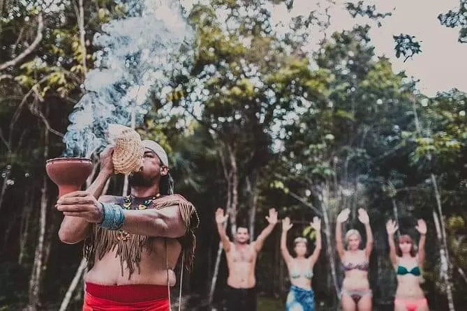 foto de un temazcal. se ve un hombre en primer plano con un intstrumento maya. atras se ve la gente pasticipando. estan en la selva