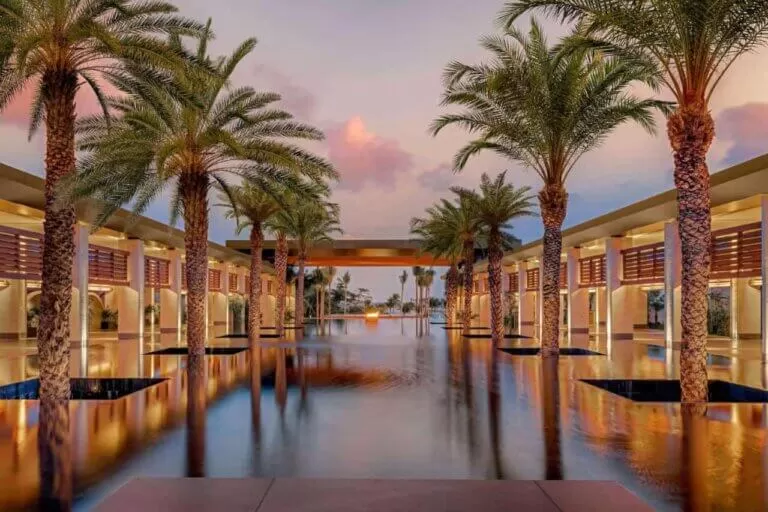 foto del hotel Conrad Tulum Riviera Maya. es el atardecer. se ve la pileta con palmeras a los costados y columnas que nacen desde la pileta donde se encuentran las habitaciones.