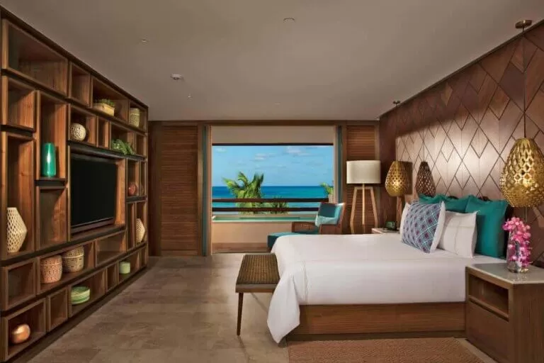 foto de la habitacion del hotel Secrets Maroma Beach Riviera. se ve una cama, con un balcon que da al mar. todo muy lujoso. paredes de madera y un gran tele al frente de la cama