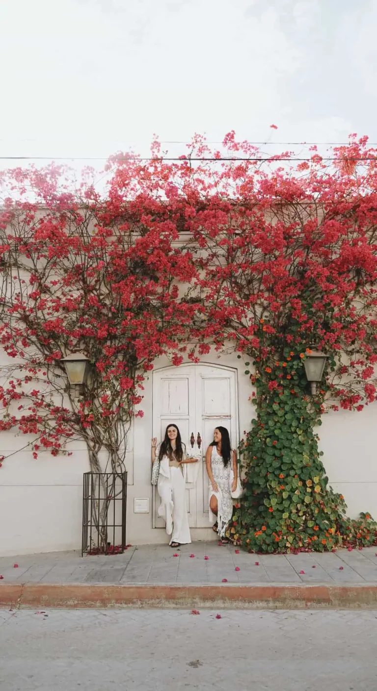 yo y pau apoyadas en una fachada de una casa en todos santos. alrededor hay una santa rita llena de flores rosas