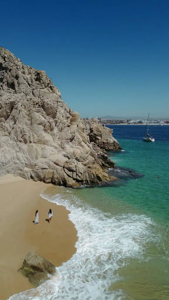 Vista aerea de la Playa del Amor, con sus aguas cristalinas y formaciones rocosas dramáticas, solo accesible por mar, y situada junto al famoso Arco de Cabo San Lucas. se nos ve a nosotras dos caminando en la playa
