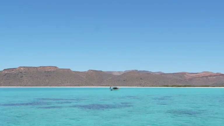 foto del mar de la isla de espiritu santo en la paz. detras se ve un barco justo en el centro del horizonte. detras se ven montañas. el agua es muy turquesa