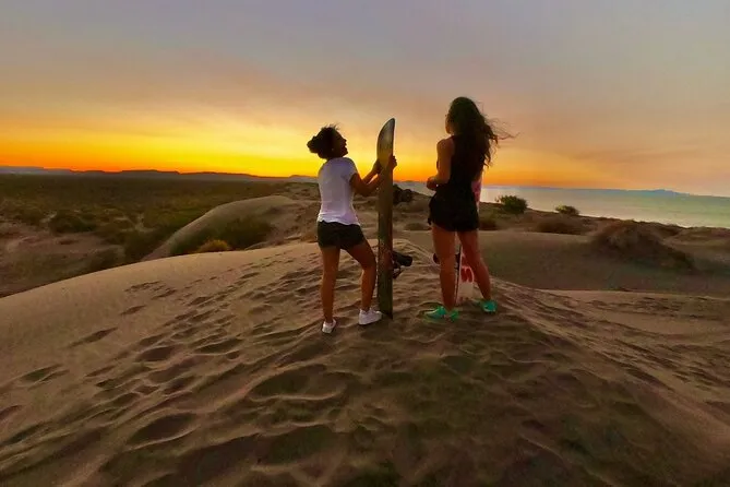 dos chicas con una board en el medio del desierto con un atardecer de fondo